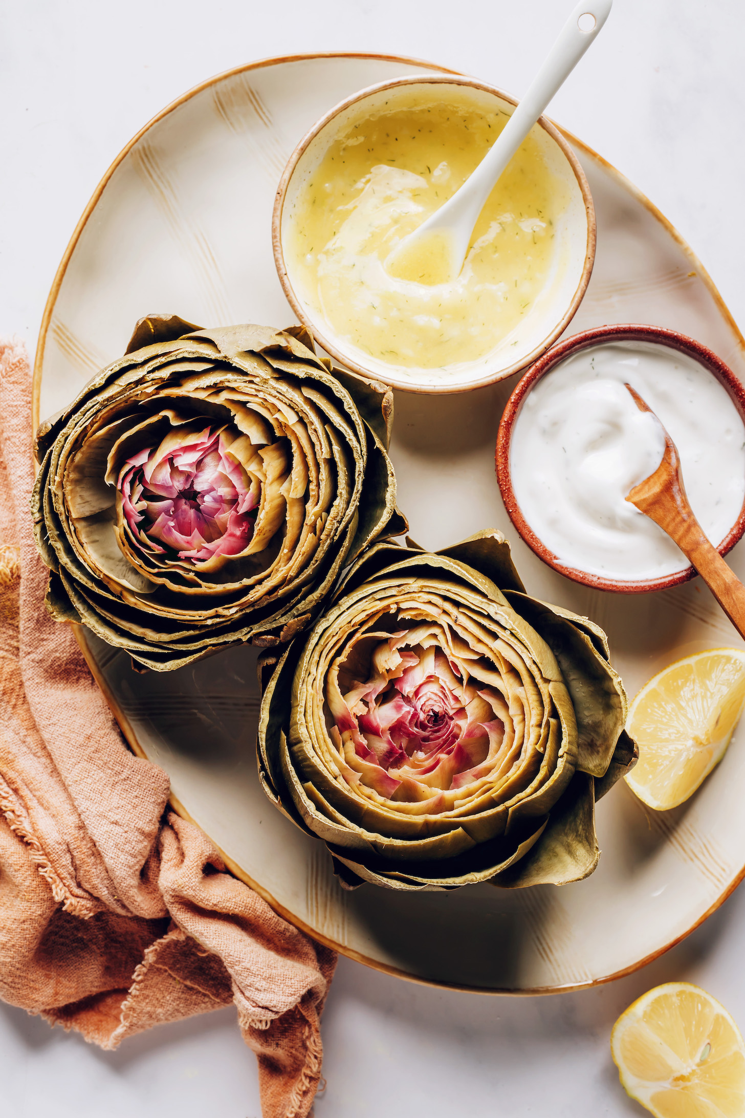 Platter with lemon wedges, steamed artichokes, and two creamy lemon dill sauces for dipping