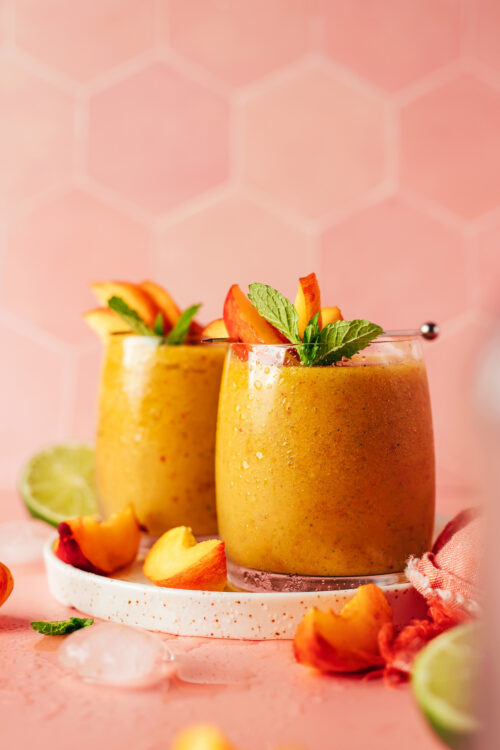 Two glasses of our frozen peach daiquiri with sliced peaches and mint resting at the top of the glasses