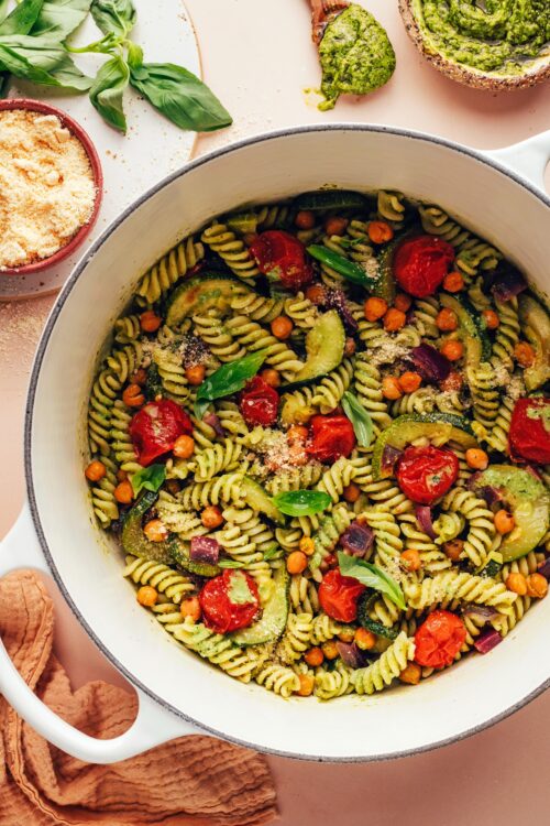 Dutch oven of zucchini pesto pasta with fresh basil, pesto, and vegan parmesan around it